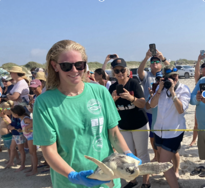 Blond woman wearing green shirt smiles while holding sea turtle in front of public crowd.
